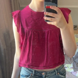 Mörkröd blus med volangdetaljer - Säljer en mörkröd blus med volangdetaljer vid axlarna och lös passform. Blusen är ärmlös och har fina sömmar upptill som ger en extra detalj. Perfekt att styla med jeans eller kjol.