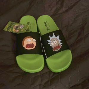 Rick and Morty sandaler grön/svart - Coola sandaler i neon-grönt med svarta remmar och tryck av Rick och Morty på ovansidan. Perfekta för dig som älskar serien och vill sticka ut med färgglada och bekväma slides. Tillverkade i syntetmaterial och har platt sula.