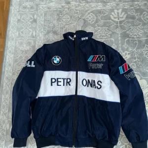 BMW Petronas F1 Jacka  - Mörkblå och vit racinginspirerad vindjacka från BMW Petronas med broderade loggor och detaljer på bröst, ärmar och rygg. Jackan har hög krage, dragkedja framtill och muddar vid ärmslut och midja. Perfekt för dig som gillar motorsport och streetwear.