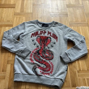 Grå Philipp Plein sweatshirt med orm - Snygg grå sweatshirt från Philipp Plein med ett stort rött och svart ormtryck och coola detaljer framtill. Tröjan har rund hals, långa ärmar och ribbade muddar. Perfekt för dig som gillar streetstyle och statement-plagg.