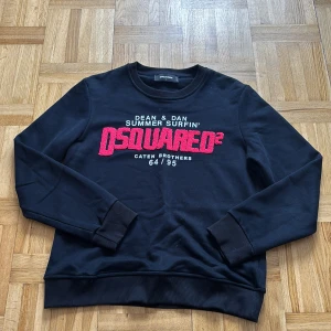 Svart sweatshirt från Dsquared2 - Svart sweatshirt från Dsquared2 med stort rött broderat logotyptryck på bröstet och vita textdetaljer. Klassisk rund halsringning, ribbade muddar och nederkant. Tillverkad i mjuk bomull, perfekt för en chill och stilren look.