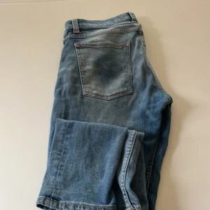 Blå jeans från Nudie Jeans Co - Säljer ett par klassiska blå jeans från Nudie Jeans Co med snygga kontrastsömmar och ikonisk våg på bakfickan. Jeansen har normal passform och är tillverkade i slitstarkt bomullsjeans. Perfekta för dig som gillar en tidlös och avslappnad look.