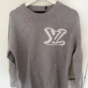 Grå stickad tröja Louis Vuitton - Snygg grå stickad tröja från Louis Vuitton med stort vitt LV-logo på bröstet. Tröjan har rund halsringning, ribbade muddar och lång ärm. Perfekt för dig som vill ha en exklusiv och stilren look med en touch av lyx.