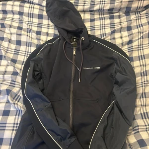 Mörkblå Porsche x Boss vindjacka - Snygg mörkblå vindjacka från Porsche x Boss med huva och dragkedja. Jackan har vita detaljer längs ärmarna och logga på bröstet. Perfekt för dig som gillar sportig stil och vill sticka ut med exklusivt samarbete.