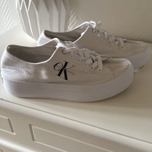 Vita sneakers från Calvin Klein Jeans - Snygga vita sneakers från Calvin Klein Jeans med tydlig CK-logga på sidan och bak. Skorna har en rejäl plattformsula och är tillverkade i canvas med vita snören. Perfekta för dig som vill ha en clean och trendig look.