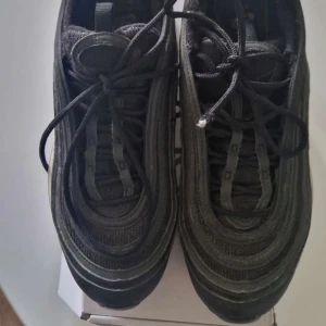 Svarta Nike Air Max 97 sneakers - Säljer ett par svarta Nike Air Max 97 sneakers med ikoniska vågformade detaljer och synlig luftbubbla i sulan. Skorna har snörning och en platt sula, tillverkade i en mix av syntet och mesh för en sportig look. Du får 5 procent på hela köpet om du även köper min Air Jordans som också finns utannonserade.