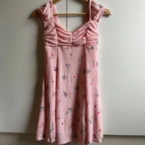 Rosa blommig kortklänning med volang - En gullig, rosa miniklänning i ett lite strukturerat tyg med blommor i olika färger på. Klänningen har rynkade detaljer vid bysten. Den är i mycket bra skick, förutom en fläck på insidan som är svart/grå (se bild 2). Fläcken syns dock inte alls från utsidan.💕