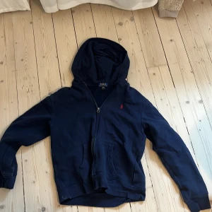 Marinblå hoodie från Polo Ralph Lauren - Snygg marinblå hoodie från Polo Ralph Lauren med dragkedja och klassisk röd broderad logga på bröstet. Tröjan har huva, kängurufickor och är tillverkad i mjuk bomull. 