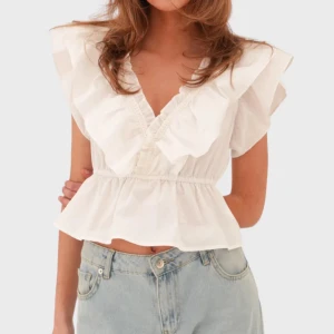 Vit blus ”June” från meet me there - Supersöt vit blus från meet me there med v-ringning och volanger längs axlar och hals. Modellen är croppad med resår i midjan för en snygg passform. Perfekt till jeans eller kjol för en fräsch look. Luftigt bomullsmaterial som passar perfekt till varmare dagar.