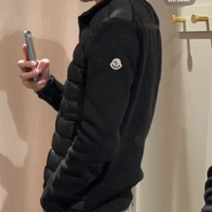 Svart cardigan från Moncler - Säljer en svart cardigan från Moncler med dragkedja framtill och quiltad design. Jackan har långärmade ärmar och en klassisk Moncler-logga på ena ärmen. Perfekt för kyligare dagar.