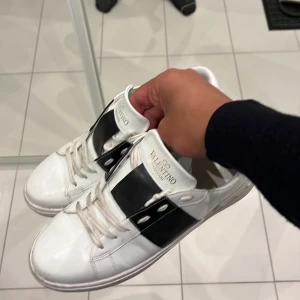 Valentino Open sneakers i vitt och svart - Säljer ett par vita Valentino Open sneakers med svart band över mitten och vita snören. Skorna har rund tå och platt sula, samt diskret Valentino-logga på plösen. Klassisk och stilren design som passar till många outfits. Har ej box eller dust bag har blivit för små försöker bli av med dom 