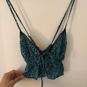 Blommig croppad linnetopp från Bershka - Supersöt blommig linnetopp från Bershka i blått och svart med tunna axelband och spetsdetaljer. Toppen har knyt framtill och är croppad, perfekt för varma dagar eller lager på lager.