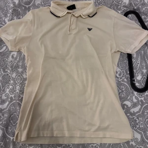 Beige pikétröja från Emporio Armani - Säljer en klassisk vit pikétröja från Emporio Armani med korta ärmar och liten logga på bröstet. Tröjan har krage med svarta och vita ränder och knäppning vid halsen. Perfekt för en stilren look.