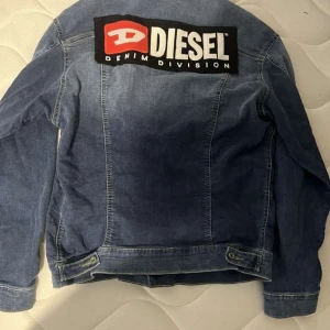 Blå jeansjacka från Diesel - Säljer en klassisk blå jeansjacka från Diesel med stort logotyp-tryck på ryggen. Jackan har knäppning framtill, två bröstfickor med lock och knapp samt klassisk krage. Perfekt för dig som gillar streetwear och denim.