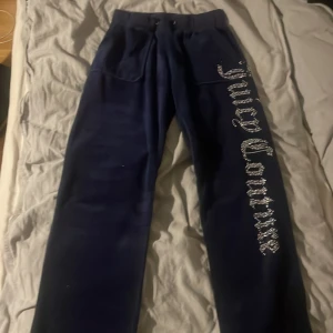 Mörkblå velourbyxor och kofta från Juicy Couture - Mörkblå mjukisset i velour från Juicy Couture med stora strasstenar som bildar loggan längs ena benet samt på ryggen av koftan. Byxorna har normal passform, hög midja och fickor både fram och bak. Koftan har fickor och luva. Perfekta för en avslappnad och trendig look.