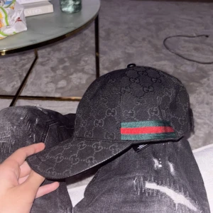 Svart Gucci keps med GG-mönster - Säljer en svart keps från Gucci i storlek M. Kepsen har klassiskt GG-monogram över hela ytan och en ikonisk grön-röd rand på sidan. Tillverkad i bomull och polyester, justerbar baktill. Perfekt för dig som vill ha en lyxig och stilren accessoar.