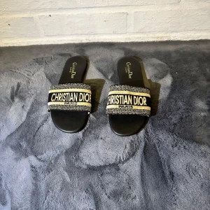 Christian Dior svarta sandaler - Svarta sandaler från Christian Dior med bred broderad rem i svart och beige med tydlig logga. Platt sula och öppen tå, perfekt för sommardagar. Lyxig känsla med snygg kontrast mellan färgerna och exklusiv brodyrdetalj.
