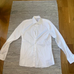 Vit skjorta från Bläck - Klassisk vit skjorta från Bläck i bomull. Skjortan har lång ärm, knappar framtill och en stilren krage med diskret mörk piping på insidan. Perfekt för en clean och stilren look.