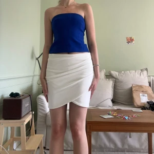 Vit omlottkjol - Säljer en vit kort omlottkjol med draperad framsida. Kjolen har en stilren look och stängs med dragkedja bak. Perfekt för dig som gillar minimalistisk stil och vill ha något enkelt men snyggt.