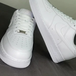Nike Air Force 1 helvita sneakers - Klassiska helvita Nike Air Force 1 sneakers med snörning och rund tå. Skorna är i skinn och har en platt sula. Perfekta för dig som gillar en clean och tidlös stil. Ikonisk Nike-logga på sidan och metallbricka vid snörningen.