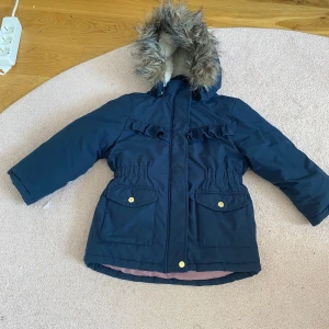 Mörkblå barn vinterjacka med fuskpäls från Name It  - Mörkblå vinterjacka från Name It med avtagbar huva och fuskpäls. Jackan har dragkedja, knappar, två stora fickor framtill och rynkade detaljer vid bröstet. Insidan är ljusrosa och fodrad för extra värme.