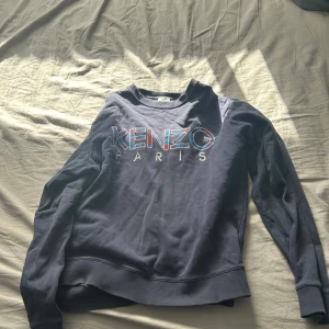 Mörkblå sweatshirt från Kenzo Paris - Säljer en mörkblå sweatshirt från Kenzo Paris med stort tryck på bröstet. Tröjan har rund halsringning, långa ärmar och ribbade muddar vid ärmslut och nederkant. Perfekt för en stilren och avslappnad look.