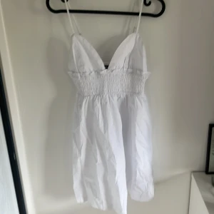 Vit kort klänning med smock från Sister - Söt vit kortklänning från Sister med smala axelband och smockad överdel. Klänningen har v-ringning och är ärmlös, perfekt för varma dagar. Lätt och luftig modell som passar till sommarens alla tillfällen.
