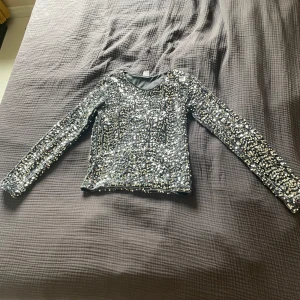 Glittrig långärmad topp från Lindex - Säljer en superglittrig långärmad topp från Lindex i storlek 170. Hela tröjan är täckt av silvriga paljetter som verkligen fångar ljuset. Perfekt för dig som vill sticka ut och glänsa lite extra!🤍🩶