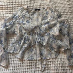 Blommig transparent blus från SHEIN - Säljer en superfin transparent blus från SHEIN med blått och vitt blommönster. Blusen har långa ärmar, volangdetaljer och knyt framtill. Perfekt för dig som gillar romantisk och luftig stil.