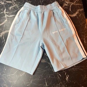 Ljusblå shorts från Palm Angels med vita ränder - Snygga ljusblå shorts från Palm Angels med vita sidoränder och logga framtill. Shortsen har elastisk midja med snörning och sidofickor. Perfekta för en avslappnad och sportig stil.