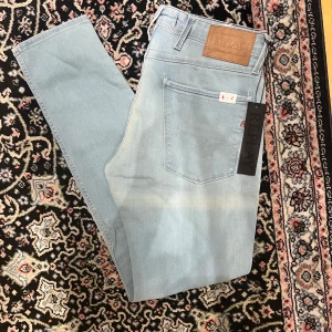 Replay Anbass 32/32 helt nya ljusblå - Säljer ett par ljusblå Replay Anbass jeans med klassisk femficksdesign och läderpatch bak i midjan. Jeansen har normal passform och är tillverkade i bomull med en liten mängd stretch för extra komfort. Perfekta för en avslappnad stil.
