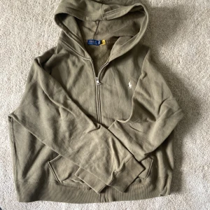 Olivgrön hoodie från Polo Ralph Lauren - Olivgrön hoodie med dragkedja från Polo Ralph Lauren. Tröjan har huva, två fickor framtill och den klassiska broderade loggan i vitt på bröstet. Mjuk insida och ribbade muddar vid ärmslut och nederkant.