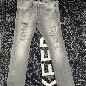 Replay jeans  - Säljer ett par grå jeans från Replay med slitna detaljer och hål på båda benen. Byxorna har fem fickor, raka ben och klassisk knappgylf. Märkeslapp bak i svart läder och diskret röd Replay-logga på bakfickan.
