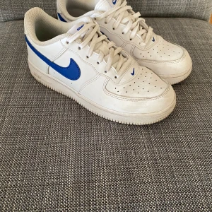 Nike Air Force 1 med blå swoosh - Säljer ett par klassiska Nike Air Force 1 sneakers i vitt skinn med blå swoosh och detaljer. Skorna har snörning, rund tå och platt sula. Perfekta för dig som gillar en tidlös och sportig stil.