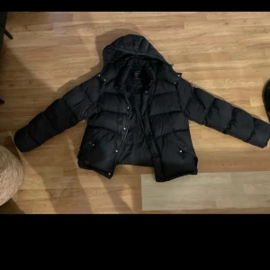 Svart pufferjacka från Morris R Club - Svart pufferjacka från Morris R Club med huva och snygga detaljer som logga på ärmen. Jackan är vadderad och har dragkedja samt knappar framtill. Perfekt för kalla dagar och riktigt skön att bära. Klassisk och stilren design som funkar till det mesta.