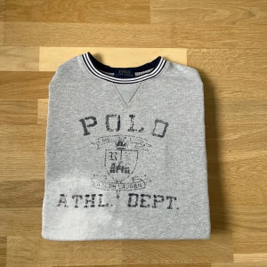 Grå sweatshirt från Polo Ralph Lauren - Grå sweatshirt från Polo Ralph Lauren med tryckt logga och text framtill. Tröjan har rund hals med randig ribbad kant i vitt och marinblått. Klassisk och enkel stil, perfekt till jeans eller joggers. Mjuk bomullsblandning för skön känsla.
