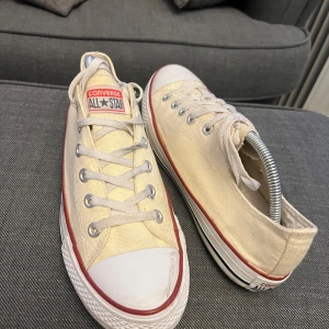 Beige Converse All Star sneakers - Säljer ett par klassiska beige Converse All Star sneakers med snörning och vit gummisula. Skorna har den ikoniska röda randen runt sulan och är tillverkade i textil med Converse-loggan på plösen.
