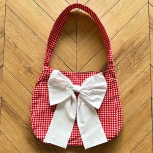 Röd och vit rutig handväska med rosett - Väska i röd och vit rutig design med en stor vit rosett framtill. Väskan har ett bekvämt, lite bredare, handtag för enkel bärning. Knappt använd. Från Motel Rocks, nypris: £28, ca 375kr +115kr i frakt. 