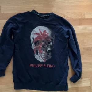  tröja från Philipp Plein - Snygg mörkblå tröja från Philipp Plein med ett stort dödskalletryck i rött och vitt på framsidan. Tröjan har långa ärmar och en rund halsringning. Perfekt för en cool och avslappnad stil.