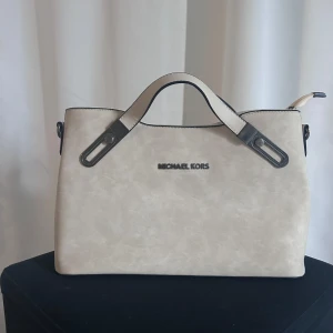 Beige handväska från Michael Kors - Snygg beige handväska från Michael Kors med svarta detaljer och logotyp framtill. Väskan har en praktisk dragkedja på baksidan och två handtag för enkel bärning. Axelband följer med 