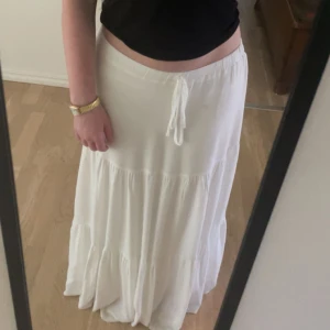 Vit långkjol med knytband - Säljer en vacker vit långkjol med ”rosett” . Kjolen har en luftig och flödande design som passar perfekt för varma dagar. Den är enkel och stilren, vilket gör den lätt att matcha med olika toppar.