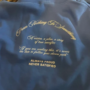 Blå hoodie från Hoodrich - Säljer en blå hoodie från Hoodrich med texten 'From Nothing To Something' och 'Always Proud Never Satisfied' tryckt på ryggen. Framtill finns en stor ficka och texten 'Hoodrich' tryckt i vitt. Perfekt för en avslappnad stil. Finns byxor till hoodien det ser man på bilden 