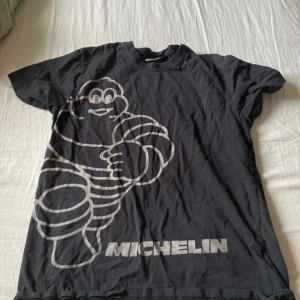 Svart t-shirt med Michelin-tryck - Cool svart t-shirt med stort vitt tryck av Michelin-mannen på framsidan. Perfekt för dig som gillar klassiska bilteman. T-shirten har en avslappnad passform och är kortärmad.