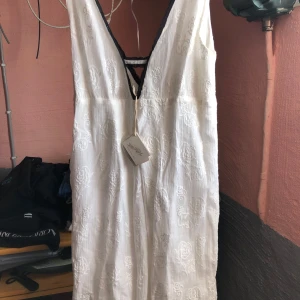 Vit klänning med broderad detalj - Elegant vit klänning med djup v-ringning och broderade detaljer. Klänningen har en öppen rygg med snörning och är ärmlös, vilket ger en stilren look. Perfekt för sommarens festligheter.