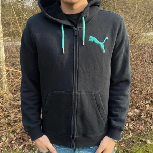 Svart hoodie från Puma - Säljer en svart hoodie från Puma med dragkedja och turkosa detaljer. Den har en stor logga på bröstet och justerbara snören i huvan. Perfekt för en avslappnad stil.