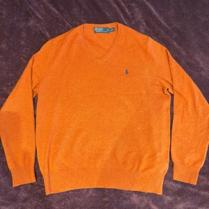 Orange tröja från Polo Ralph Lauren - Säljer en snygg orange tröja från Polo Ralph Lauren med v-ringning och broderad logga i blått på bröstet. Tröjan har långa ärmar och ribbade muddar vid ärmslut och nederkant. Perfekt för en stilren look!