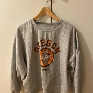 Grå sweatshirt med tryck - Säljer en grå sweatshirt med tryck från Oregon Sports Association. Tröjan har en rund halsringning och långa ärmar. Perfekt för en avslappnad stil.