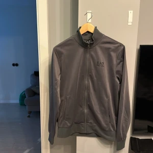 Grå kofta från EA7 - Snygg grå jacka från EA7 med dragkedja framtill och hög krage. Jackan har långa ärmar och ribbade muddar vid ärmslut och nederkant. Perfekt för en stilren look.