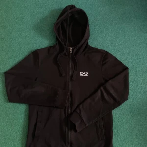 Svart huvtröja från Emporio Armani - Säljer en stilren svart huvtröja från Emporio Armani med dragkedja och EA7-logga på bröstet. Tröjan har långa ärmar och en praktisk huva med snörning. Perfekt för en casual look. Storleken är S men passar även som M.