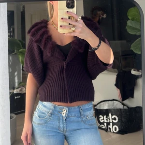 Stickad kofta - Säljer en mörklila kofta med fuskpälsdetaljer längs kragen. Koftan är ribbad och har en unik design med kortare ärmar. Perfekt för att ge en lyxig touch till din outfit. Passar bra till både jeans och kjol.